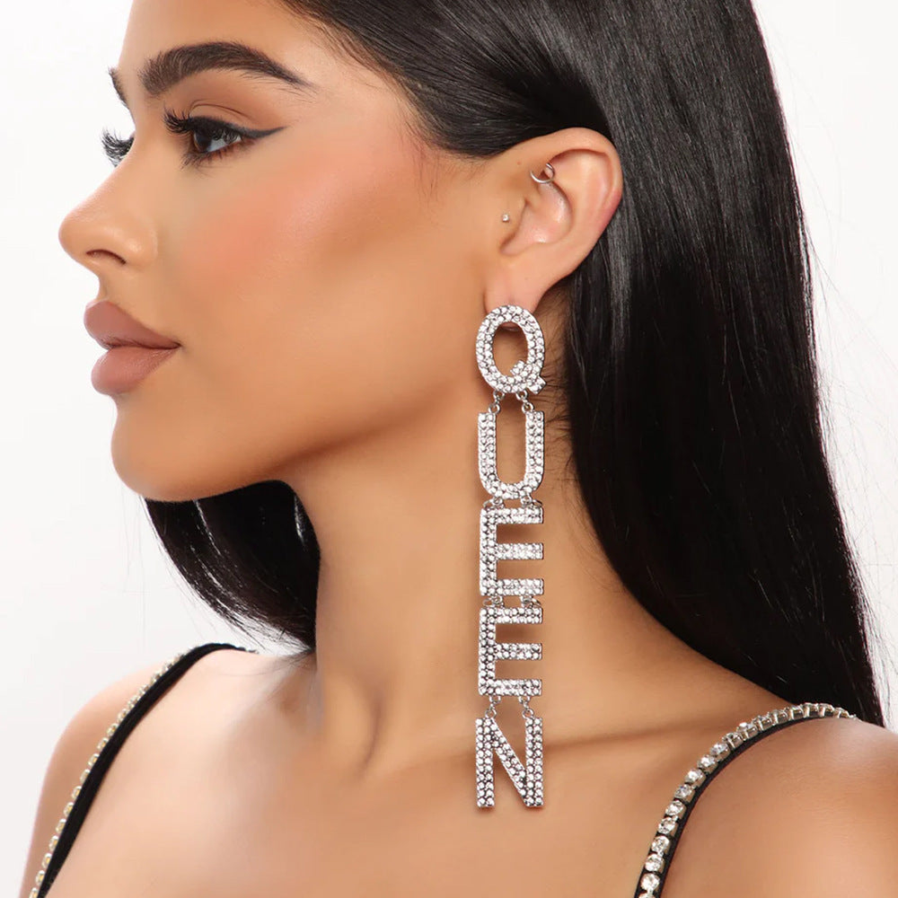 Rhinestone-embellished drop earrings spelling the word "QUEEN" displayed on cracked grey cylindrical rod against a red textured background.