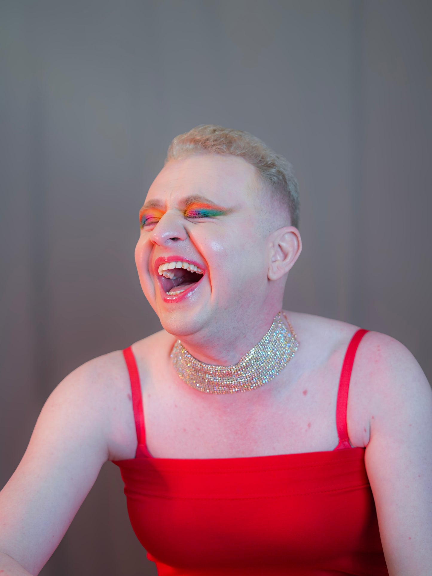 Person wearing a red dress and sparkling necklace with colorful makeup against a plain background