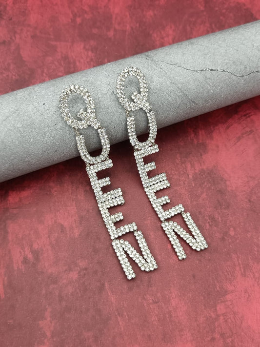 Rhinestone-embellished drop earrings spelling the word "QUEEN" displayed on cracked grey cylindrical rod against a red textured background.