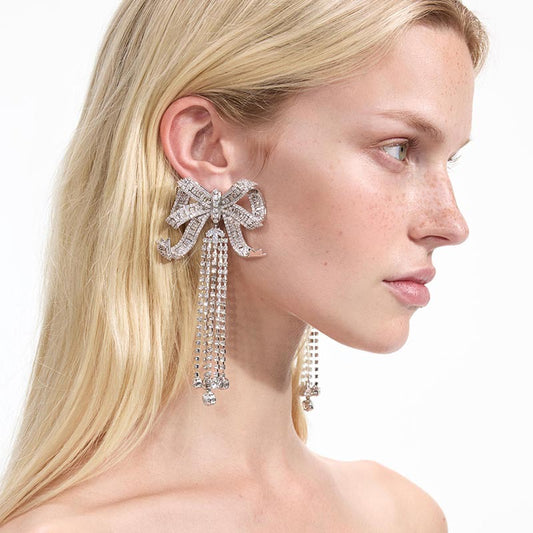 Close-up of a rhinestone bow-shaped earring with cascading strands, worn by a person with long blonde hair. A statement piece symbolizing tribute and pride.