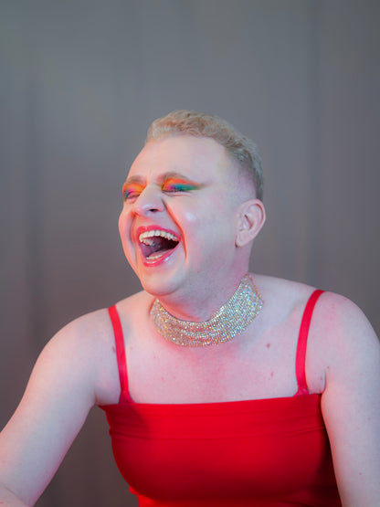 Person wearing a red dress and sparkling necklace with colorful makeup against a plain background