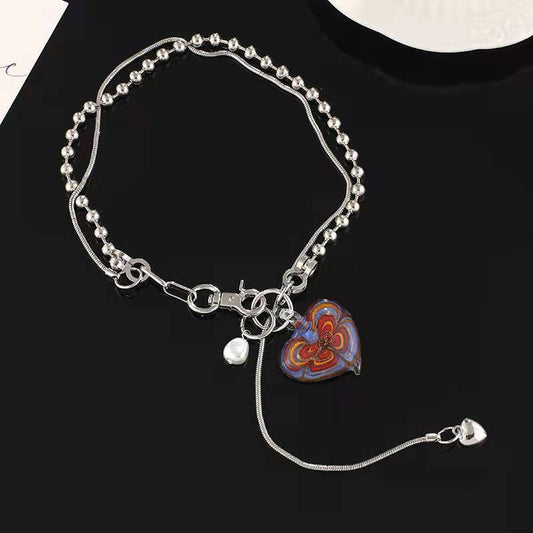 Person wearing a black V-neck top and blazer, showcasing the Nisra Heart Silver Chain – a silver necklace with a red-blue heart pendant, white accent bead, and a long drop strand ending in a small heart charm.