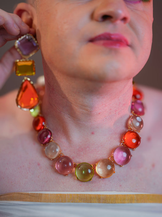 Colorful gemstone necklace and matching earrings placed on a printed page. The necklace features multicolored faceted stones in gold-toned frames, arranged symmetrically. Earrings include burgundy, purple, and amber stones in a geometric drop design.